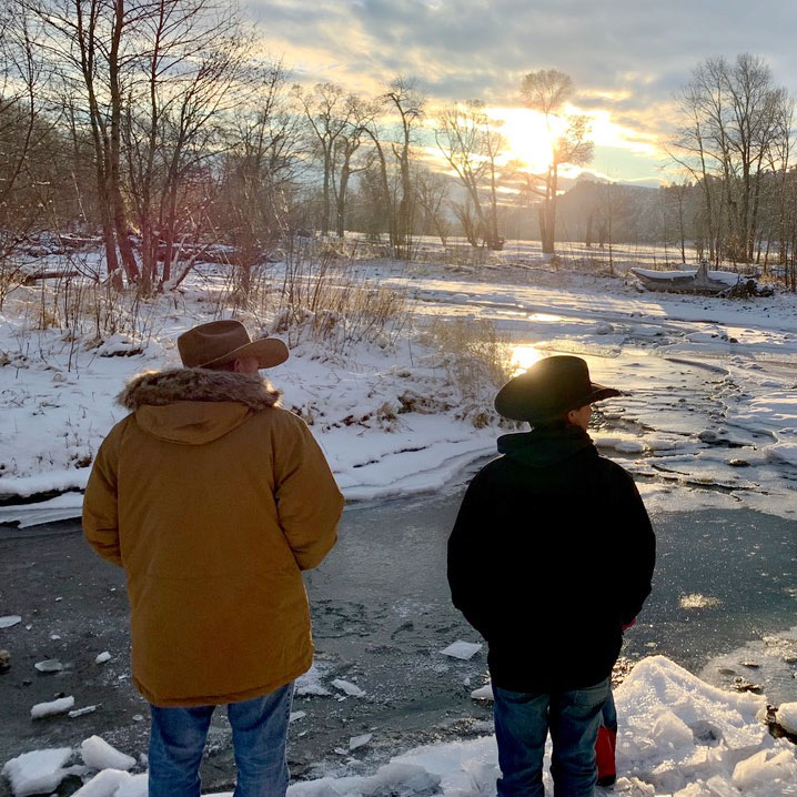 Two men standing near a river in the snow looking at a sunset